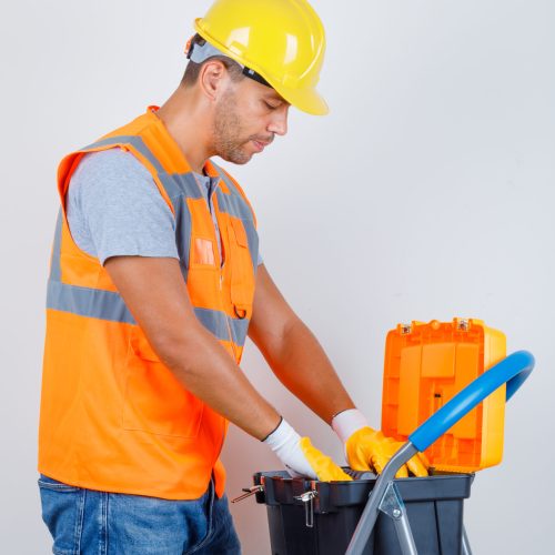 Male builder in uniform, jeans, helmet, gloves looking for something in toolbox .